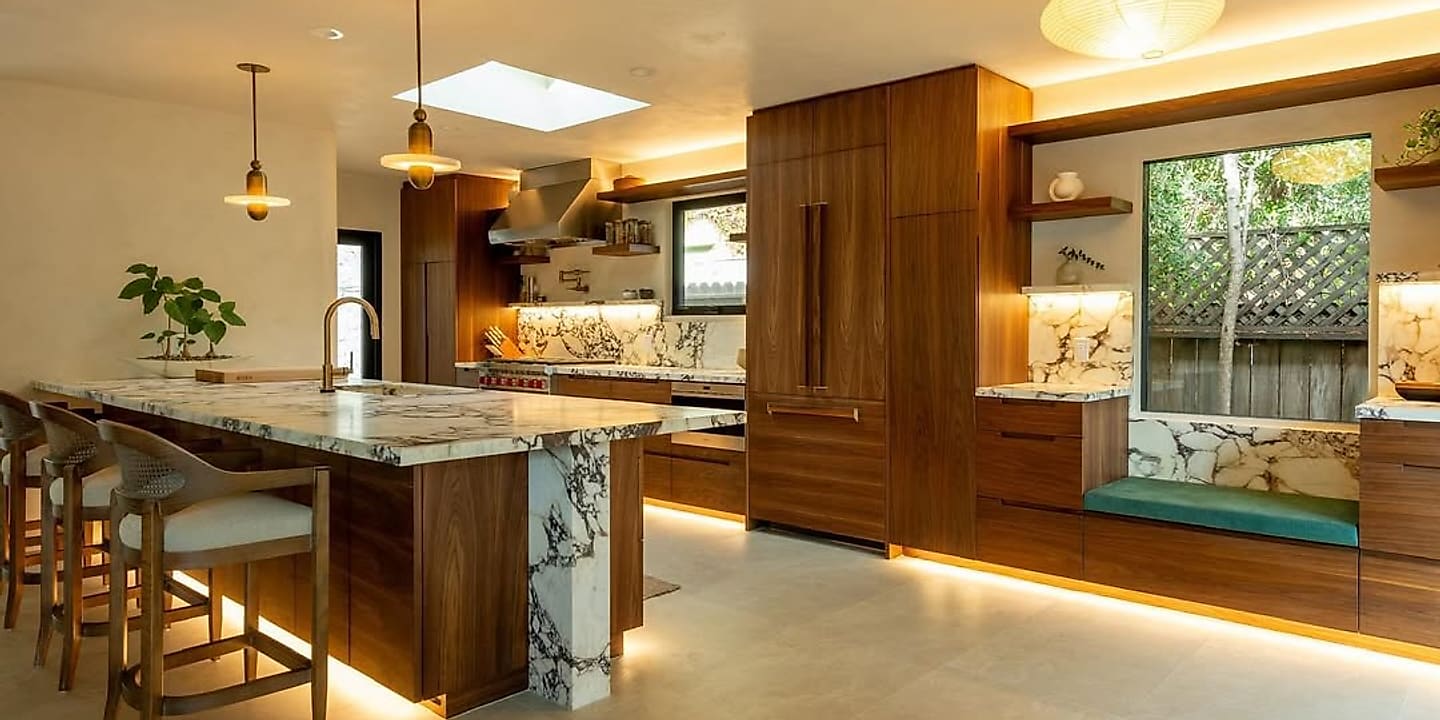 A walnut kitchen with a Calacatta stone island and backlit shelving, lit by a skylight.