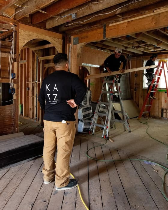 A carpenter in a Katz Construction shirt inside a house stripped back to its timber frame.