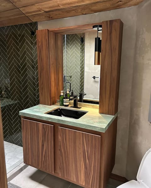 A walnut vanity with a stone counter and a custom mirror surround, against green herringbone tile.